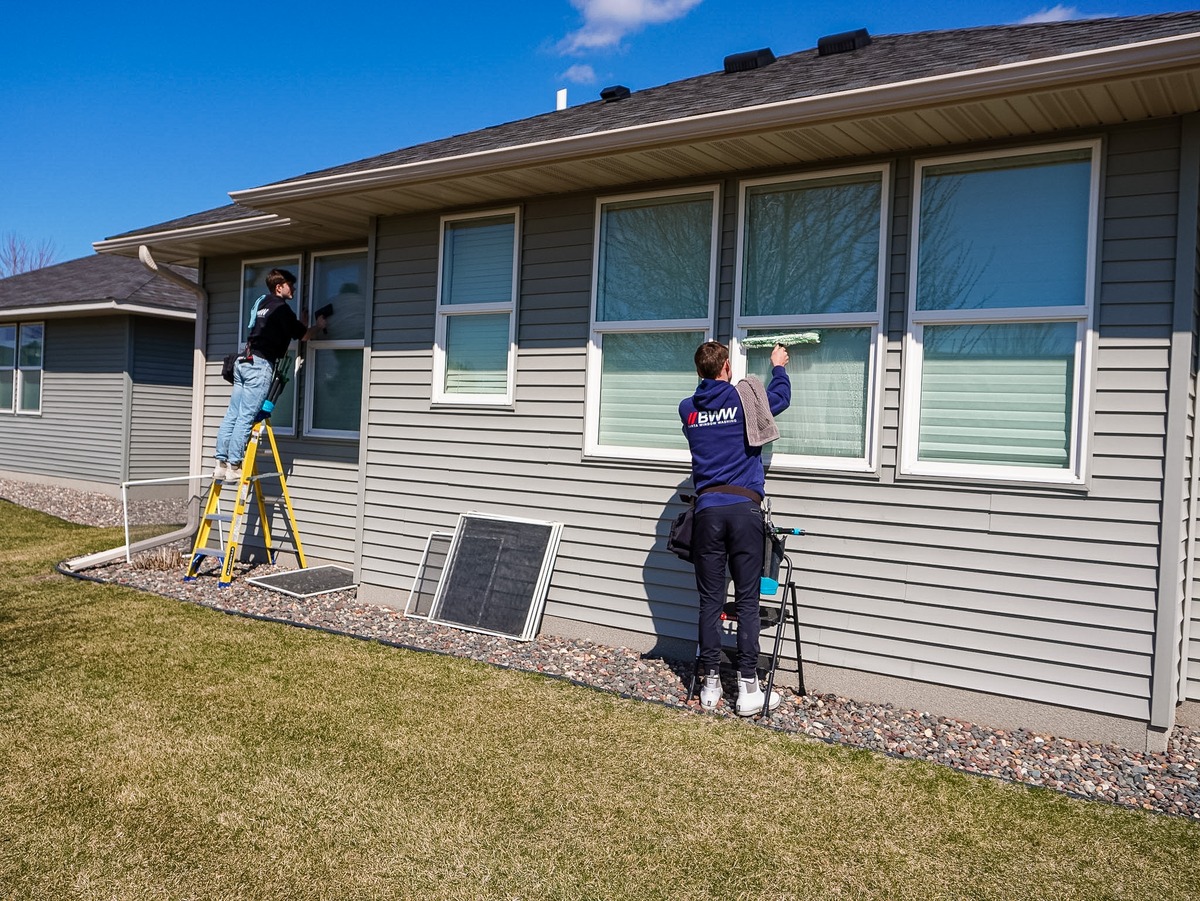 Two Barta Window Washing technicians cleaning exterior windows on a home, with screens removed nearby